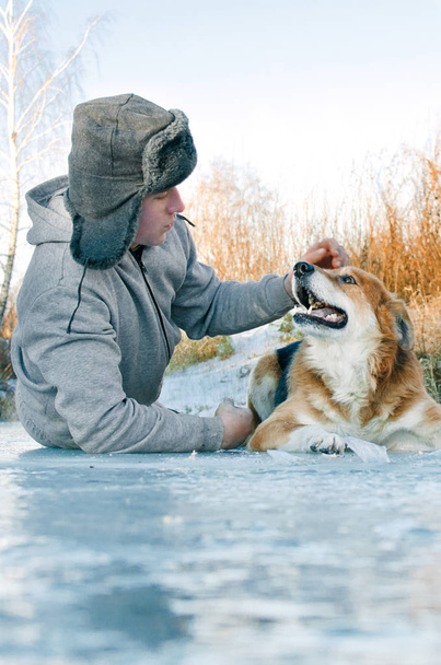 jovem em uma caminhada com um cão deitado no gelo e acariciando o cão na cabeça
 - Foto, Imagem