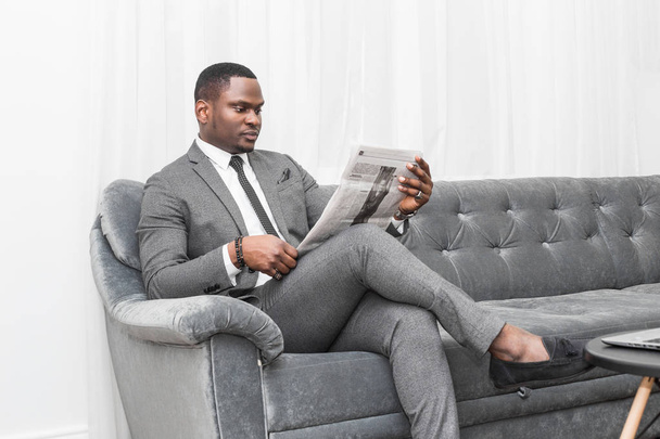 Young African American businessman in a gray suit reading a newspaper while sitting on a sofa. - Foto, afbeelding