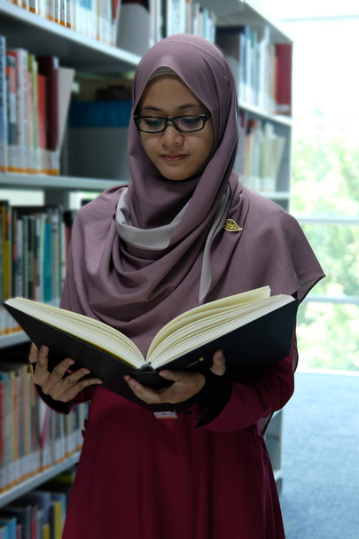 eine schöne junge Hijab-Frau liest ein Buch, nachdem sie in einer Bibliothek ein Buch aus dem Bücherregal genommen hat. - Foto, Bild