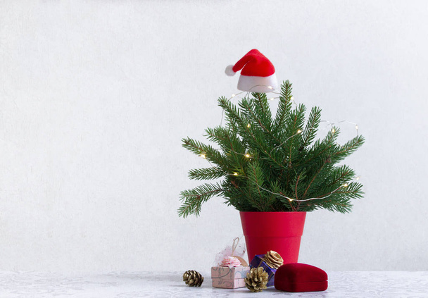 on a light background there is a Christmas tree decorated with lights in a red pot under it. Gifts of cones - Foto, imagen