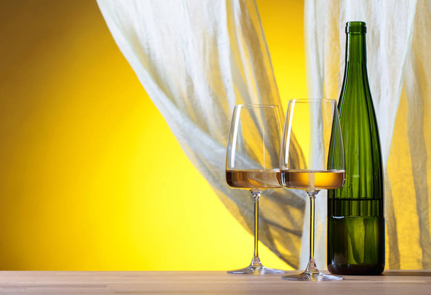 Γυαλιά και ένα μπουκάλι λευκό κρασί σε κίτρινο φόντο. Κίτρινο καθαρό ύφασμα φτερουγίζει στον άνεμο. Χώρο αντίγραφο. - Φωτογραφία, εικόνα