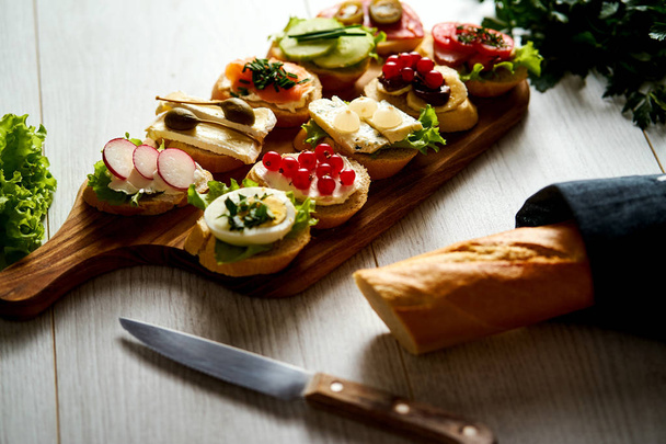 board with colorful mini sandwiches or tapas along with a knife baguette and parsley on a table - Photo, Image