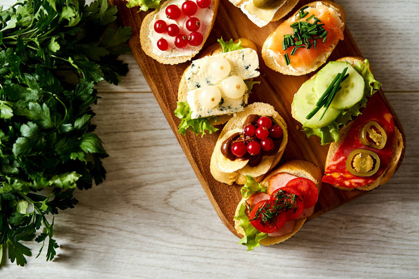 wooden board with colorful mini sandwiches or tapas along with a baguette and parsley on a light table - Photo, Image
