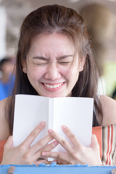 Женщина улыбается и прячет лицо за книгой, отдыхая вечером в ресторане рядом с морем и пляжем
. - Фото, изображение