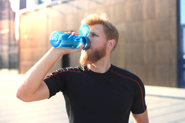 Handsome adult man drinking water from fitness bottle while standing outside, at sunset or sunrise. Runner. - Foto, immagini