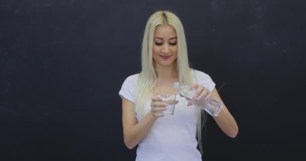 healthy blond woman pouring fresh water from a bottle into a glass and smiling on camera, over black background - Felvétel, videó
