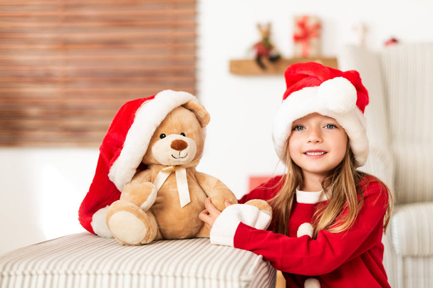 Cute young girl wearing santa hat holding her christmas present, soft toy teddy bear. Happy kid with xmas present, smiling and looking at camera. - Foto, imagen