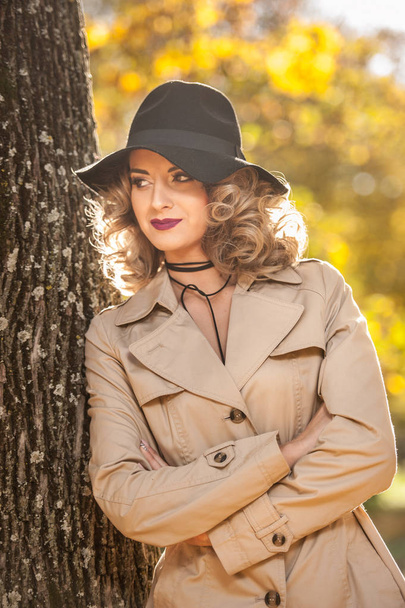 Beautiful blonde woman with cream coat, long legs and black hat in a autumn scene.  Portrait of a very beautiful, elegant and sensual woman with curly hair and sexy legs  posing in autumn park. - Zdjęcie, obraz