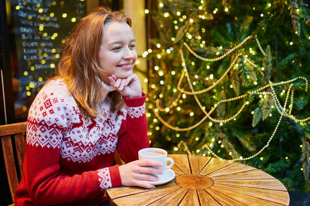 Cheerful young girl in holiday sweater drinking coffee or hot chocolate in cafe decorated for Christmas - Fotoğraf, Görsel