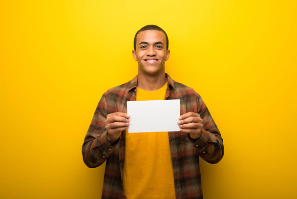 Νεαρός αφρικανική αμερικανική άνθρωπος σε ζωντανό κίτρινο φόντο κρατώντας ένα άδειο λευκό πλακάτ για την εισαγωγή μιας έννοιας - Φωτογραφία, εικόνα