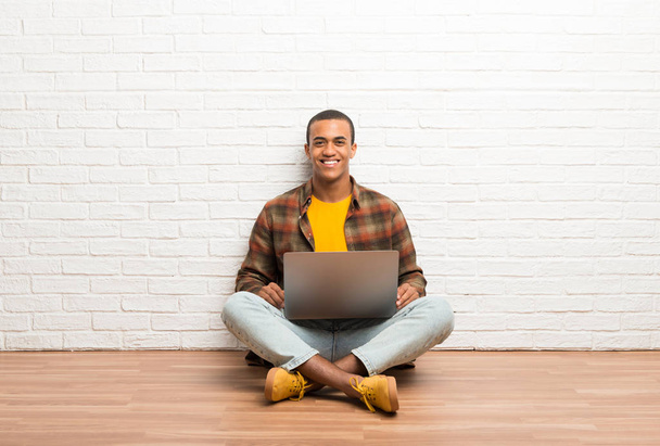 African american man sitting on the floor with his laptop posing with arms at hip and laughing looking to the front - Фото, изображение