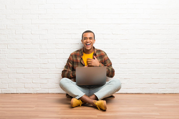 Afro-Américain assis sur le sol avec son ordinateur portable souriant beaucoup tout en mettant les mains sur la poitrine
 - Photo, image