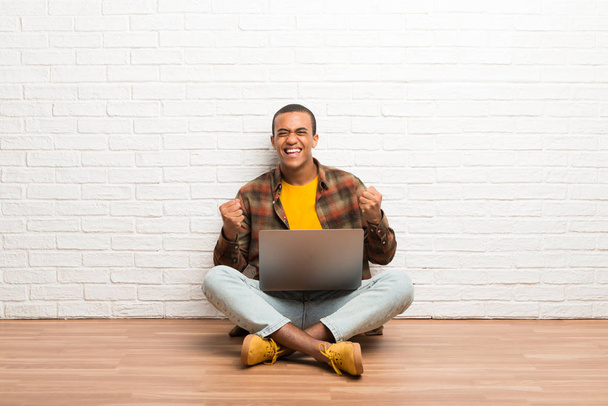 African american man sitting on the floor with his laptop celebrating a victory - Photo, Image