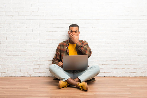 Афроамериканець людиною, сидячи на підлозі, зі своїм ноутбуком прикриття рота руками за щось недоречним - Фото, зображення
