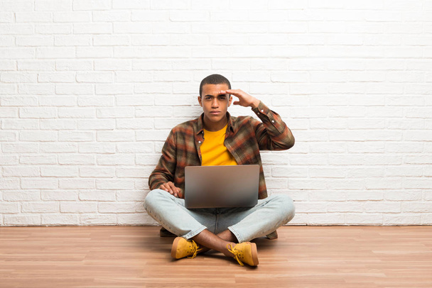 African american man sitting on the floor with his laptop looking far away with hand to look something - Photo, image