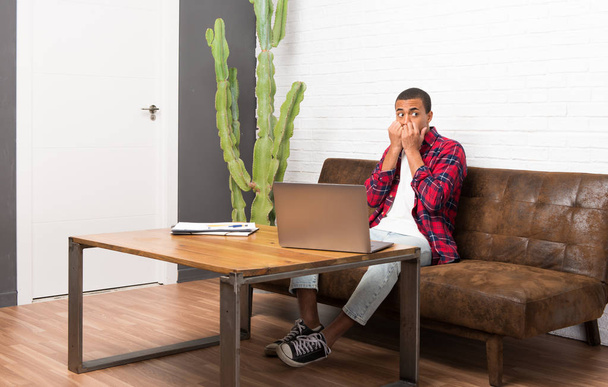 hombre afroamericano con portátil en la sala de estar es un poco nervioso y asustado poniendo las manos a la boca
 - Foto, Imagen