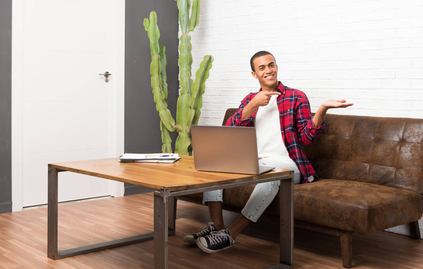 Αφρικανική αμερικανική άνθρωπος με το laptop στο χώρο του σαλονιού κρατώντας copyspace φανταστικό στην παλάμη για να εισαγάγετε μια διαφήμιση - Φωτογραφία, εικόνα