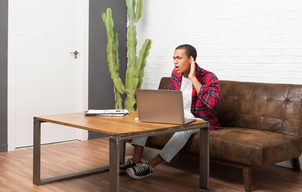 Afrikanisch-amerikanischer Mann mit Laptop im Wohnzimmer, der etwas hört, indem er die Hand ans Ohr legt - Foto, Bild
