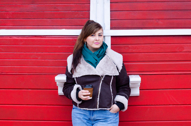 Plus size model in a winter look drinks coffee near a wooden red house. - Fotó, kép