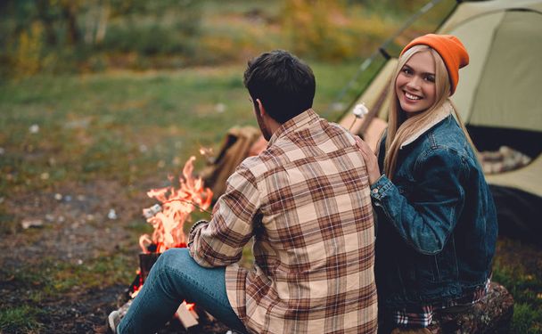 Young couple of tourists are exploring new places. Attractive woman and handsome man are spending time together on nature. Sitting near bonfire and touristic tent in forest and making marshmallow. - Valokuva, kuva