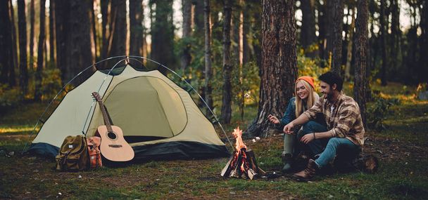 Young couple of tourists are exploring new places. Attractive woman and handsome man are spending time together on nature. Sitting near bonfire and touristic tent in forest and making marshmallow. - Photo, Image