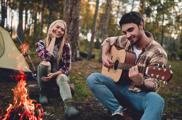 Young couple of tourists are exploring new places. Attractive woman and handsome bearded man are spending time together on nature. Sitting near bonfire and touristic tent in forest and playing guitar. - Фото, изображение