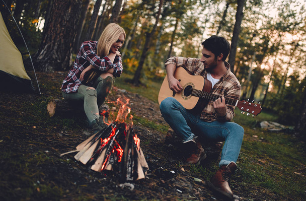Young couple of tourists are exploring new places. Attractive woman and handsome bearded man are spending time together on nature. Sitting near bonfire and touristic tent in forest and playing guitar. - Fotó, kép