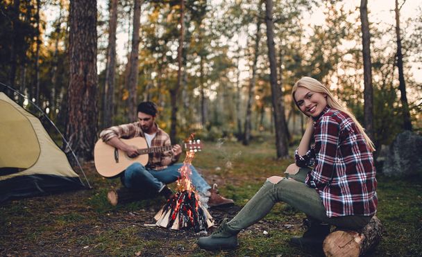 Young couple of tourists are exploring new places. Attractive woman and handsome bearded man are spending time together on nature. Sitting near bonfire and touristic tent in forest and playing guitar. - Фото, зображення