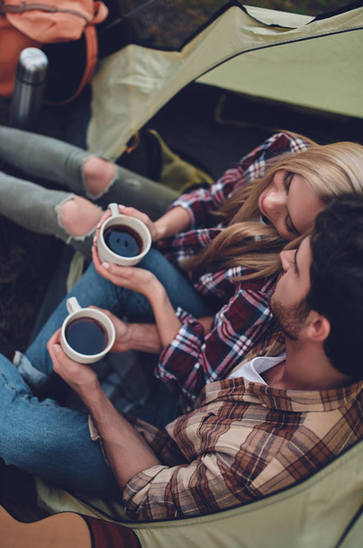 Young couple of tourists are exploring new places together. Attractive young woman and handsome bearded man are spending time together on nature. Sitting in touristic tent in forest and drinking tea. - Photo, Image