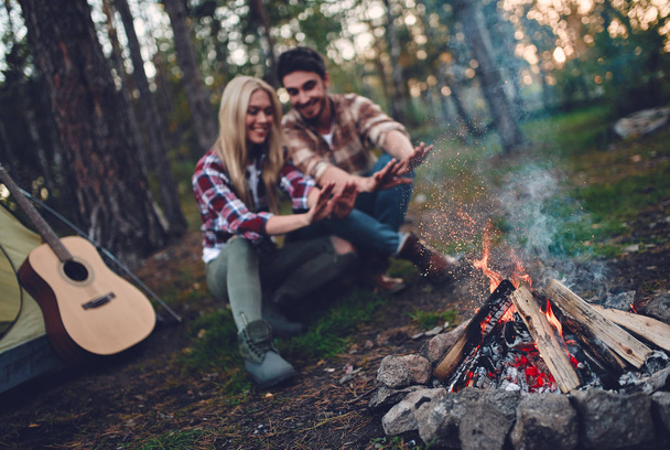 Young couple of tourists are exploring new places. Attractive young woman and handsome bearded man are spending time together on nature. Sitting near bonfire and touristic tent in forest. - Foto, immagini