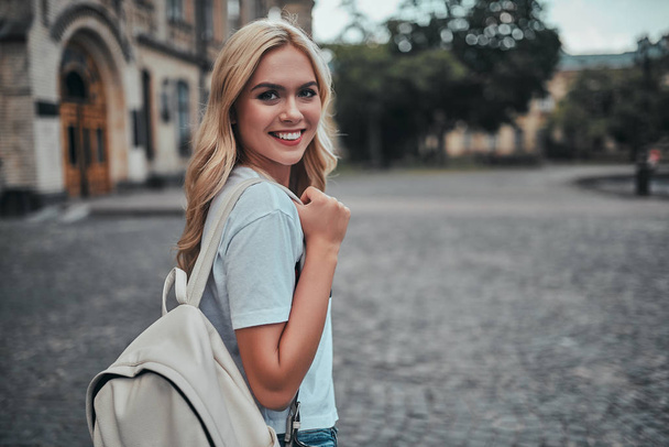 Attractive young woman is standing near university with backpack. Female student outdoors. - Фото, изображение