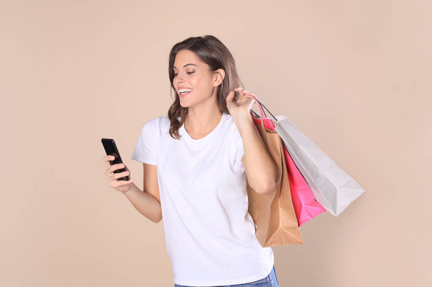 Cheerful young girl in basic clothes using mobile phone while holding shopping bags isolated over beige background. - Фото, изображение