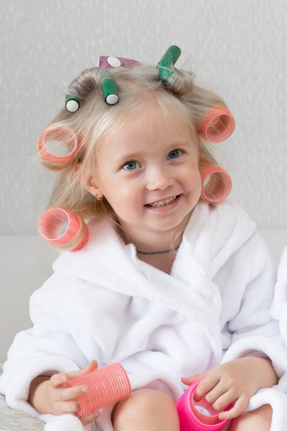 Girl blonde with curlers in her hair. Little girl in a white coat with hairpins in the hair on her head. - Valokuva, kuva