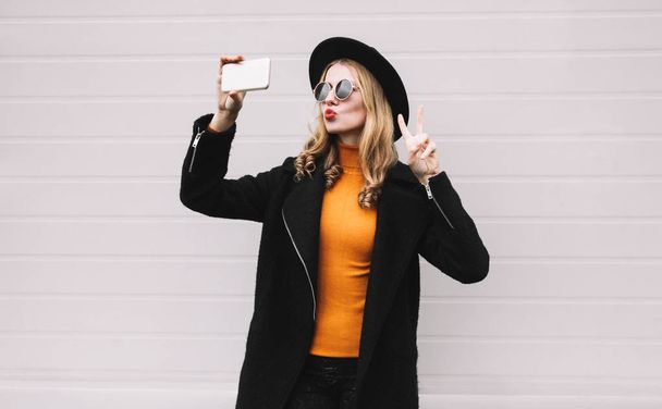 Fashion, technology and people concept - cool girl taking selfie picture by smartphone, elegant female model blowing red lips sends an air kiss posing in city on gray wall background - Foto, Imagem