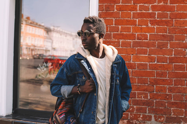 Fashion african man in jeans jacket, with backpack walking on city street, brick wall background - Foto, afbeelding