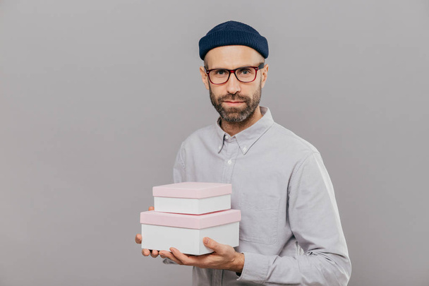 Photo of serious male model dressed in festive clothes, holds gifts in hands, going to congratulate friend with birthday, ready for party wears spectacles isolated over grey wall. This present for you - 写真・画像