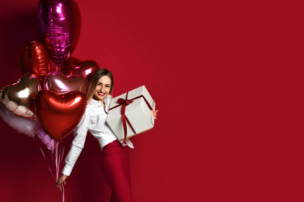Beautiful woman with present gift box and purple pink heart balloons happy smiling on red background - Photo, Image