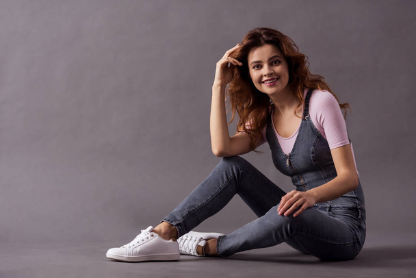 Portrait of a young beautiful woman full-length sitting on the floor on a gray background and a smile to the camera. - Fotografie, Obrázek
