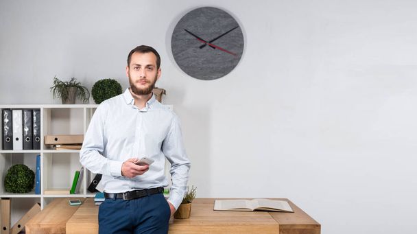 Theme office business. Handsome young caucasian man confident and strong with beard standing in bright room on working place. dressed stylishly shirt and trousers. Uses technology. Hand mobile phone. - Fotografie, Obrázek