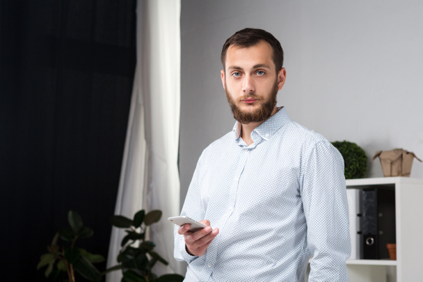 Theme office business. Handsome young caucasian man confident and strong with beard standing in bright room on working place. dressed stylishly shirt and trousers. Uses technology. Hand mobile phone. - Foto, imagen