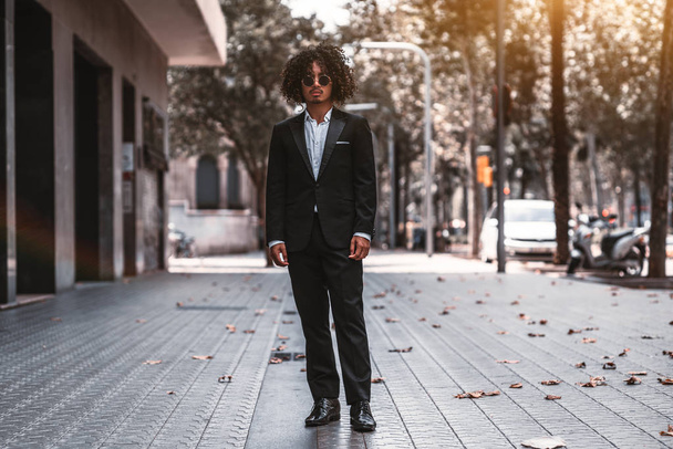 A confident Asian man entrepreneur with curly hair and in a business suit is standing in the middle of sidewalk in urban settings with buildings on the left and a road on the right - Foto, afbeelding