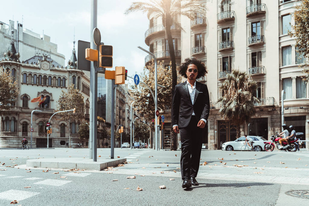 A handsome curly Asian businessman in sunglasses and a formal suit is confidently crossing the road on the crosswalk near traffic lights, with facades of buildings in the background, Barcelona, Spain - Valokuva, kuva