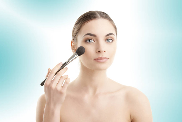 Close-up of a young woman holding brush in her hand and applying makeup. Isolated on light blue background.  - Photo, Image