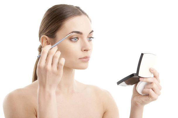 Studio shot of a beautiful young woman applying makeup to her eyebrow while standing at isolated white background and holding makeup palette in her hand. - Photo, Image