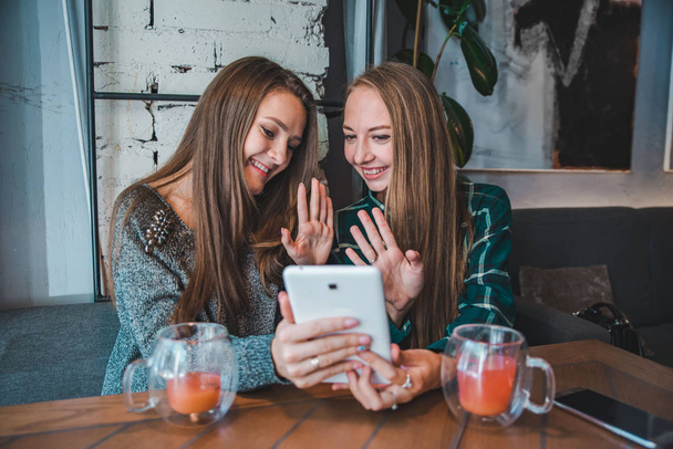 Две юные красавицы разговаривают в видеочате с помощью планшета. сидя в кафе пить фруктовый чай
 - Фото, изображение
