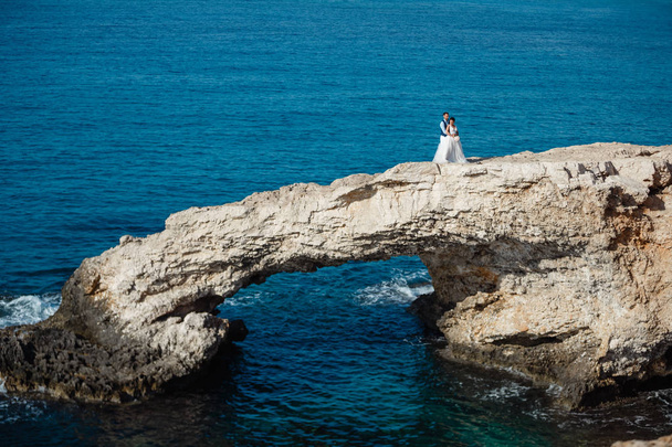 Belle jeune mariée souriante et marié marchant sur la plage, embrassant et s'amusant, cérémonie de mariage près des rochers et de la mer. Cérémonie de mariage sur la côte de Chypre
 - Photo, image