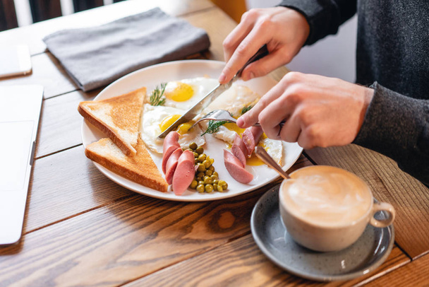 The man eat. Knife and fork in hand. Breakfast on the table.. American style breakfast with fried eggs, sausage, green peas and toast. The concept of a healthy morning. - 写真・画像