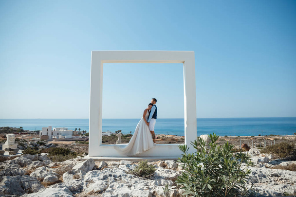 schöne lächelnde junge Braut und Bräutigam, die am Strand spazieren gehen, sich küssen und Spaß haben, Hochzeitszeremonie in der Nähe von Felsen und Meer. Hochzeitszeremonie an der Küste Zyperns - Foto, Bild