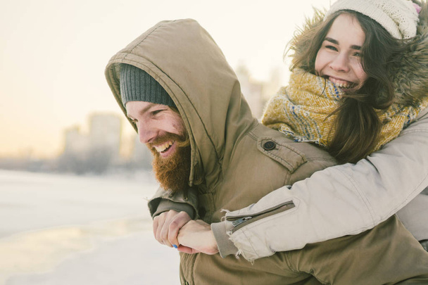 Theme New Year Christmas Mood Winter Snow Holidays Valentine Day. Young Caucasian couple lovers joy, laughter fooling water in city park. man holds woman on shoulders as backpack. - Photo, Image