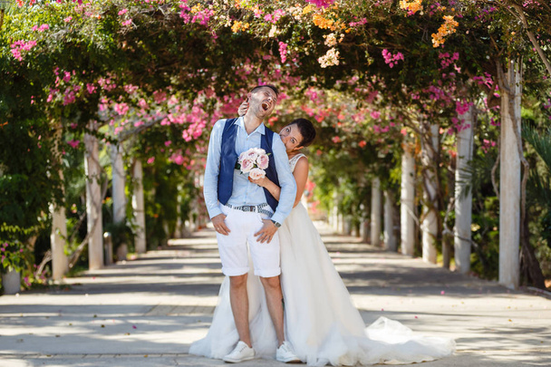 Mooie lachende jonge bruid en bruidegom lopen op het strand, kussen en plezier, huwelijksceremonie in de buurt van de rotsen en de zee. De ceremonie van het huwelijk op de kust van Cyprus - Foto, afbeelding